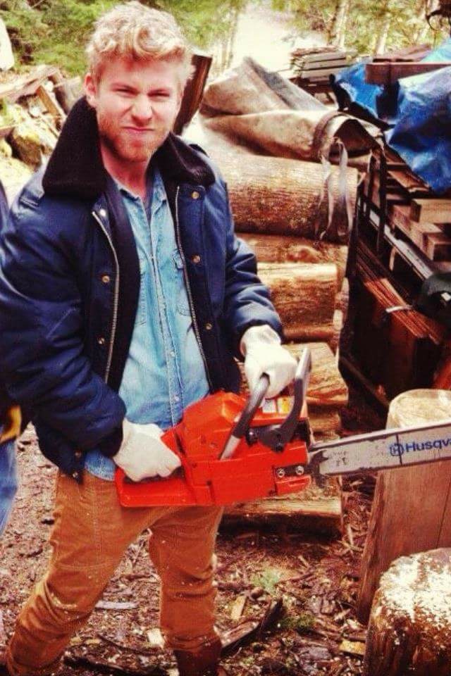 Jace Ardiel in work gloves and jacket, outdoors with a chainsaw near stacked logs